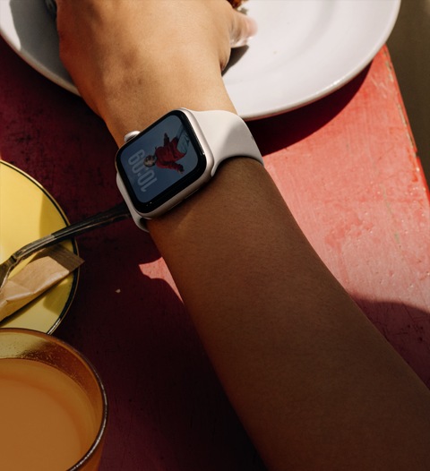 Le bras d’une personne sur la table à déjeuner, Apple Watch SE 3 sur son poignet, boîtier en aluminium comète, écran toujours allumé, portrait d’une personne sur le cadran, bracelet sport comète
