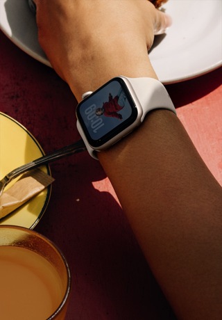 Le bras d’une personne sur la table à déjeuner, Apple Watch SE 3 sur son poignet, boîtier en aluminium comète, écran toujours allumé, portrait d’une personne sur le cadran, bracelet sport comète