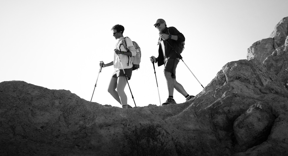 Two people wearing Apple Watch devices and hiking on a rocky trail with poles and gear